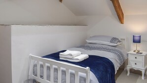 Un dormitorio con cama blanca, manta azul, mesita de noche con lámpara y vigas de madera a la vista.