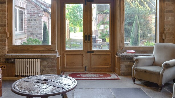 A room with a large wooden door, two windows, a radiator, a round table, and a chair. - 4 bedroom accommodation in Low Dinsdale (Low Dinsdale)