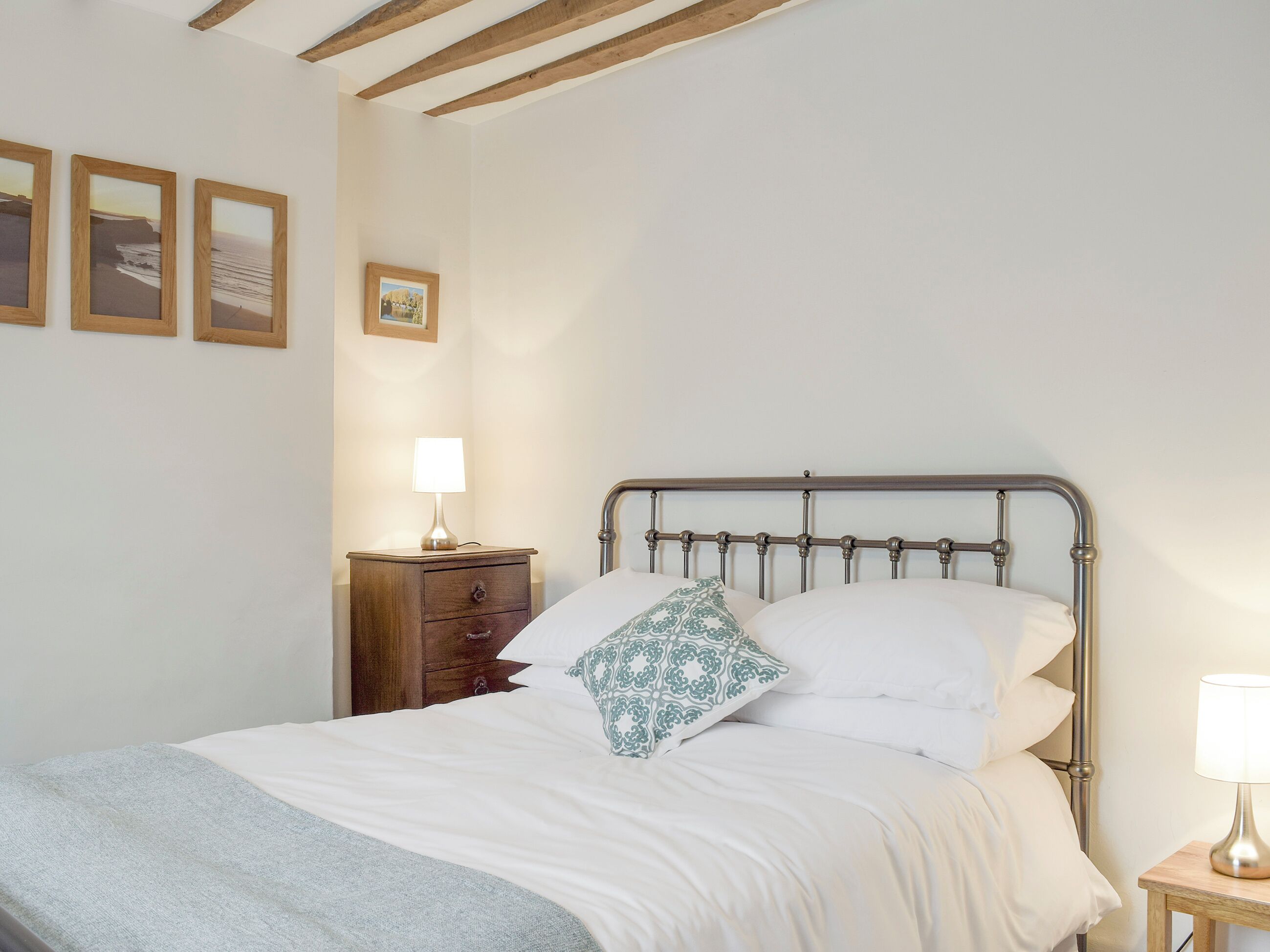 A neatly made bed with white linens and a patterned pillow, a wooden nightstand with a lamp, and framed pictures on the wall.