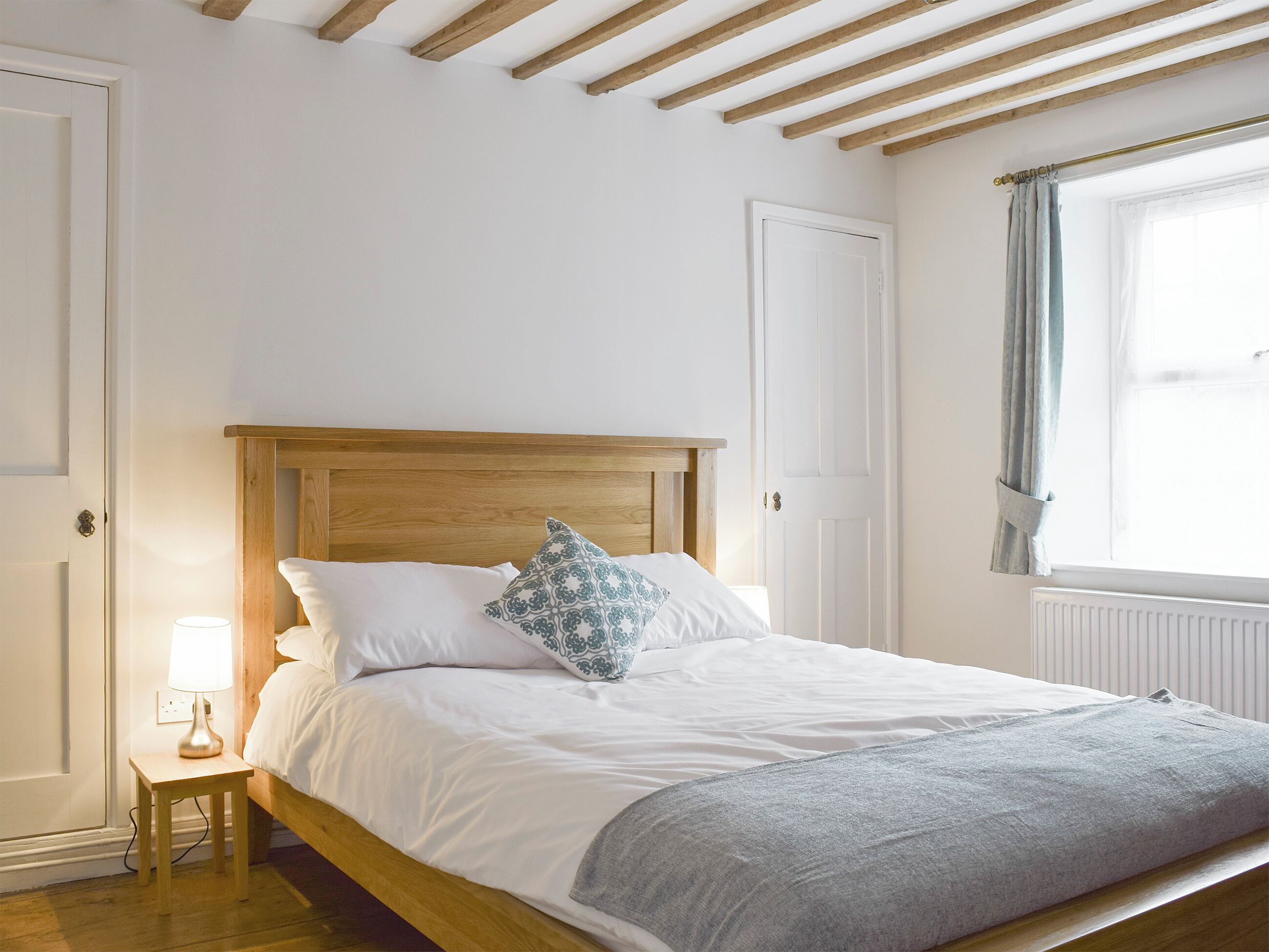 A bedroom with a wooden bed frame, white bedding, a bedside lamp, and a window with curtains.