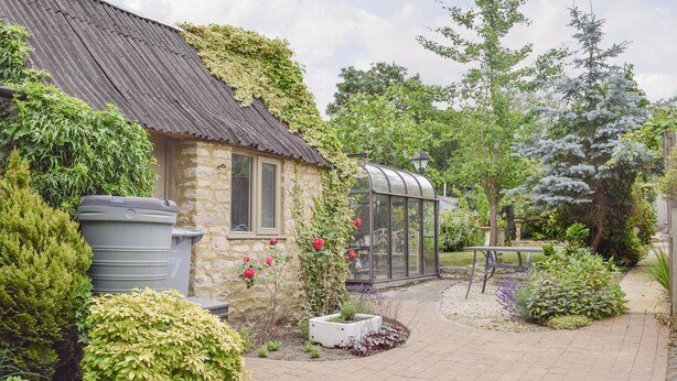 A stone house with a corrugated metal roof, a conservatory, and a garden with various plants and a pathway. - 3 bedroom accommodation in Bampton, near Witney (Bampton, near Witney)