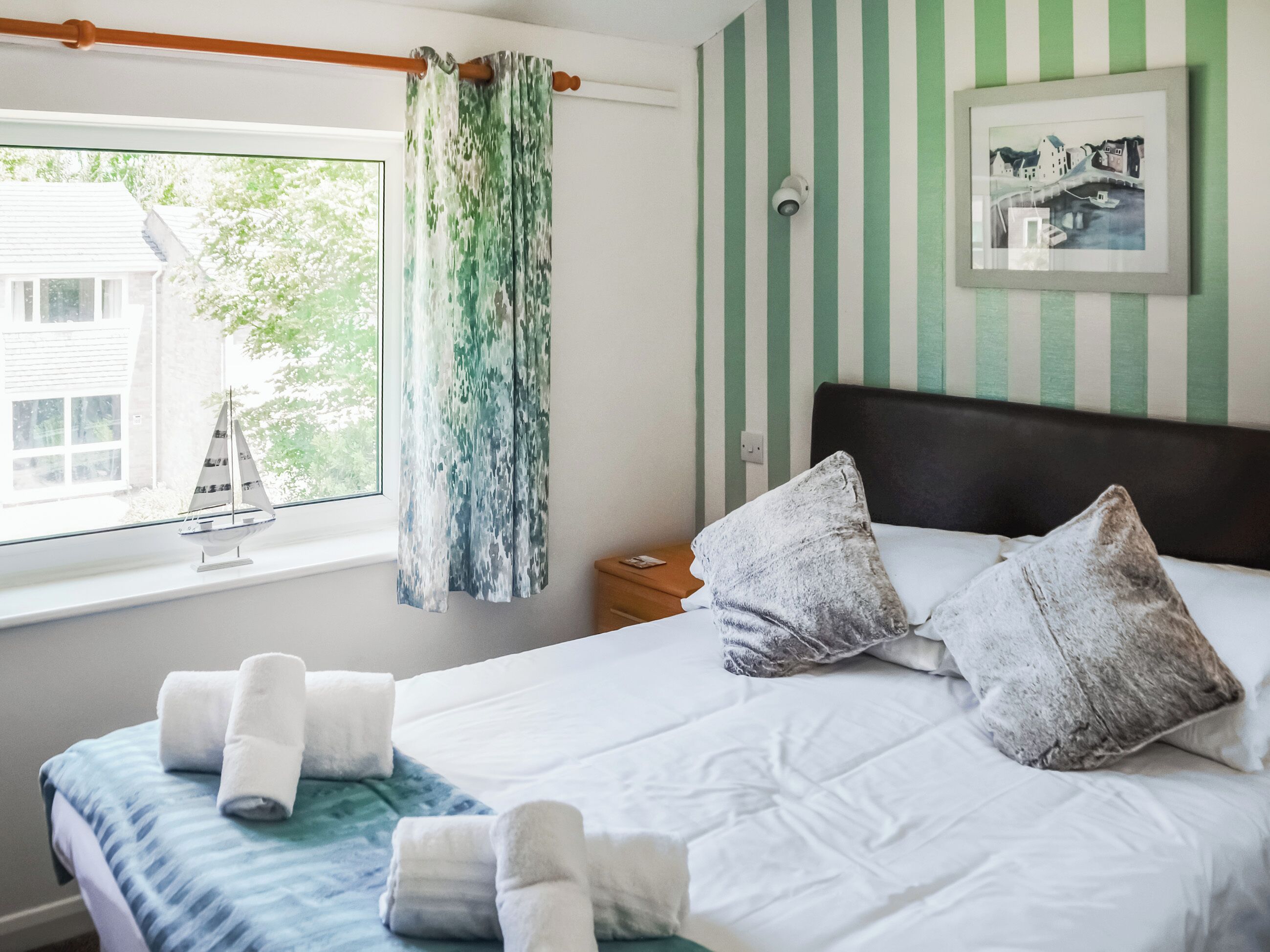 Ein Schlafzimmer mit einem Bett, Kissen, einem Fenster mit Blick auf ein Haus, einem Vorhang und einem gerahmten Bild an der Wand.