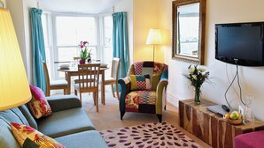 A living room with a sofa, armchair, and a dining table with chairs. A TV is mounted on the wall, and there is a window with curtains. - 2 bedroom accommodation in Bowness-on-Windermere (Bowness-on-Windermere)