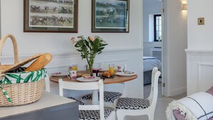 Un coin repas avec une table en bois, des chaises blanches, un panier de pain et deux tableaux historiques encadrés accrochés au mur.