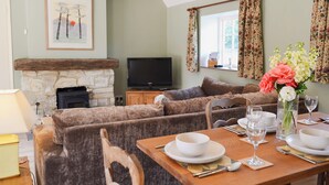 A cozy living room with a stone fireplace, a television, and a dining area set with a table, chairs, and a vase of flowers. - Cowbeech Farm Cottage (Cowbeech, near Hailsham)