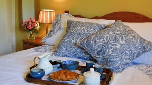 A neatly made bed with blue and white patterned pillows, a bedside table with a lamp and a vase of flowers, and a tray with croissants, coffee cups, and a teapot. - Cowbeech Farm Cottage (Cowbeech, near Hailsham)