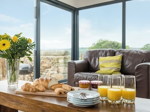 Disposição de pequeno-almoço com pastelaria, sumo e flores junto a uma janela com vista para um muro de pedra.