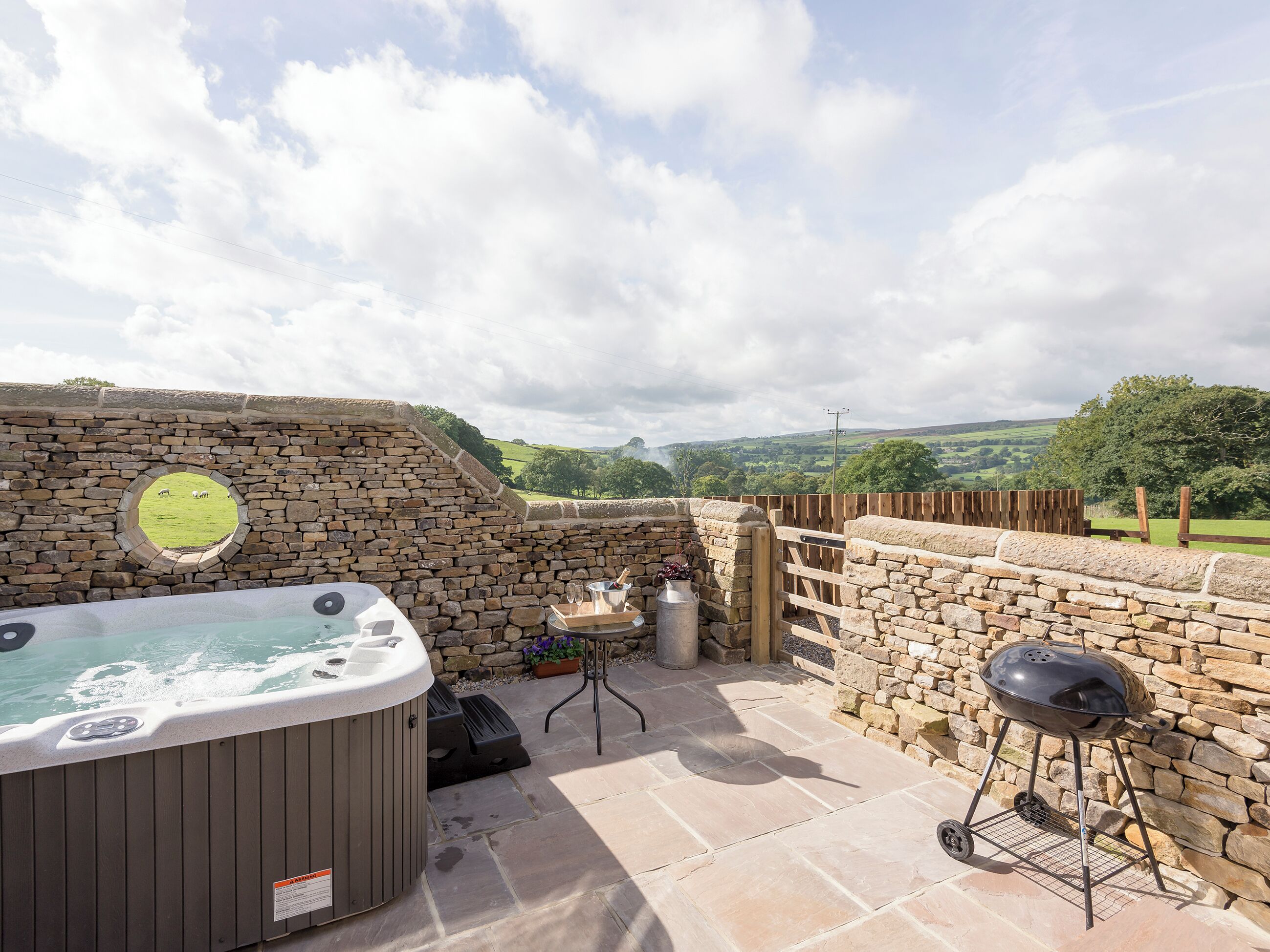 A stone wall with a hot tub, a grill, and a wooden fence overlooking a green landscape.