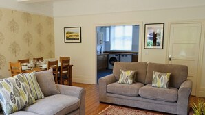 A living room with two sofas, a dining table, and chairs. There is a kitchen area visible through an open door. - 2 bedroom accommodation in Sheringham (Sheringham)