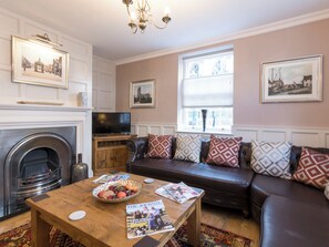 A living room with a fireplace, a television, a sofa with patterned cushions, and a table with a bowl of fruit and a magazine.