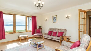 A cozy living room with a sofa, armchairs, a small table with magazines, a view of the sea through large windows, and a chandelier. - 3 bedroom accommodation in Gairloch (Gairloch)