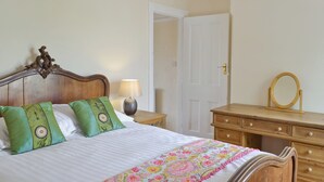 A neatly made bed with green and white pillows, a floral bedspread, and a wooden headboard. There is a wooden dresser with a mirror and a nightstand with a lamp.