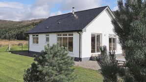 A white single-story house with a dark roof, surrounded by a grassy yard and trees, with a mountainous backdrop. - 2 bedroom accommodation in Acharacle (Acharacle)