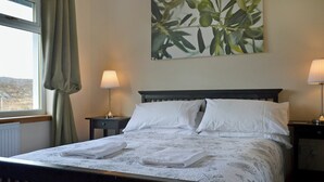 A neatly made bed with white linens and a folded towel, two bedside lamps, a window with curtains, and a wall-mounted plant. - 2 bedroom accommodation in Acharacle (Acharacle)