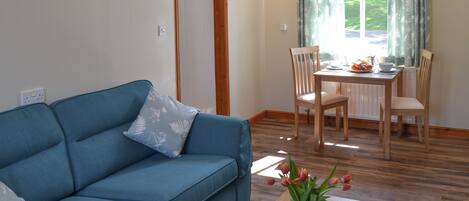 A living room with a blue sofa, a wooden dining table with chairs, a vase with flowers, and a window with curtains.