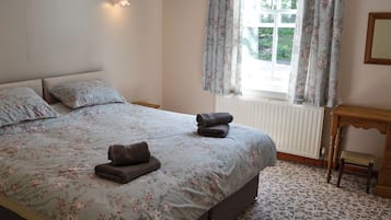 A bedroom with a bed, floral bedding, a bedside table, a window with curtains, and a radiator.