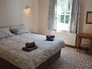 A bedroom with a bed, floral bedding, a bedside table, a window with curtains, and a radiator. - 1 bedroom accommodation in Cawston, near Norwich (Cawston, near Norwich)