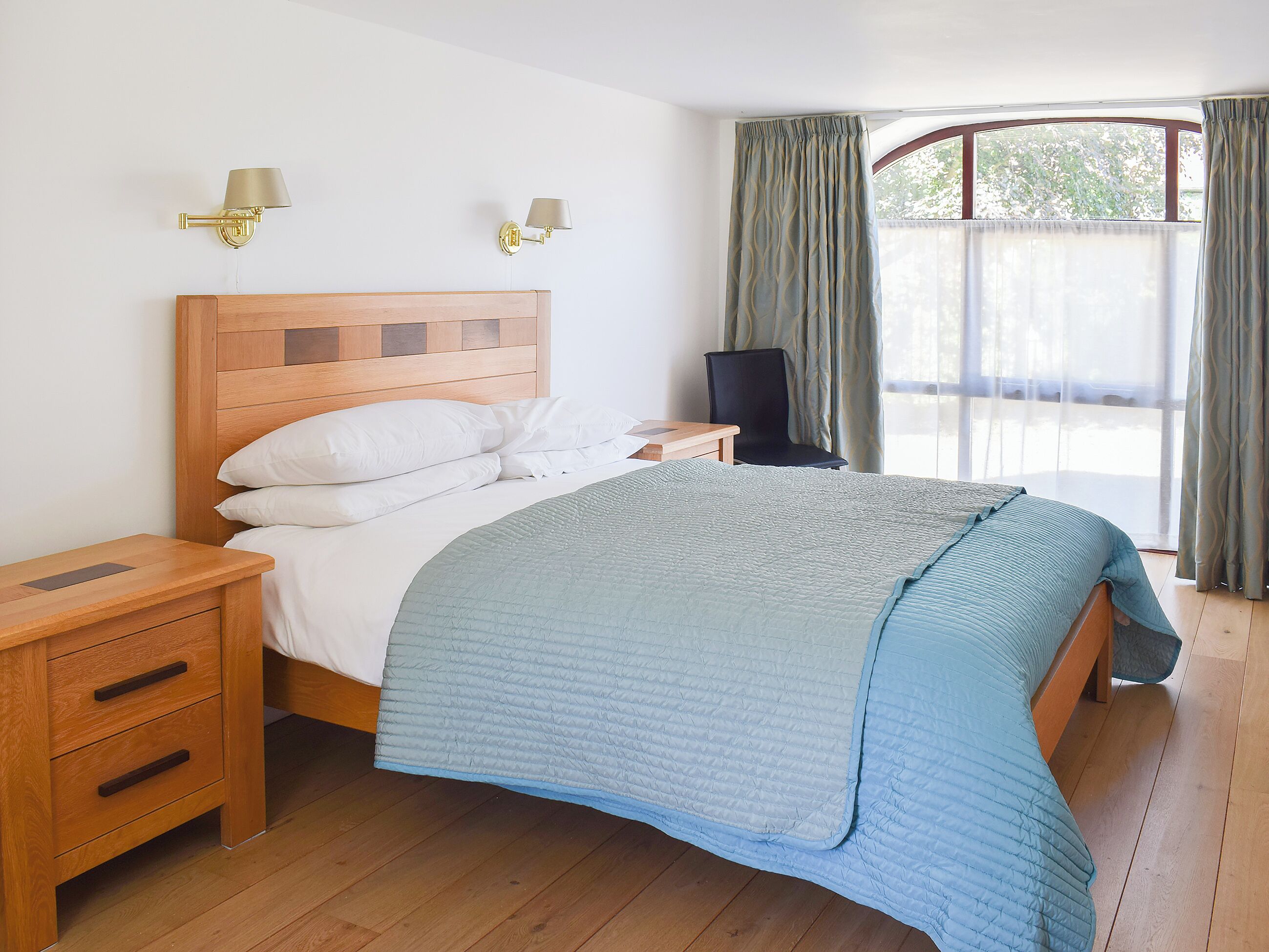 A neatly made bed with a blue quilt, wooden headboard, bedside tables with drawers, and two wall-mounted lamps.