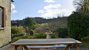 Une table de pique-nique en bois, située dans un jardin, d’où l’on peut admirer un bâtiment en briques et une colline en arrière-plan.