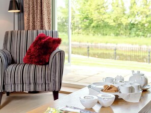 A striped armchair with a red pillow, a dining table with white ceramic dishes and cups, and a view of a lake through a large window.