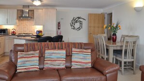 Ein Wohnzimmer mit braunem Ledersofa, einem Esstisch mit Stühlen, einer dahinterliegenden Küche und einem an der Wand hängenden Kunstwerk.