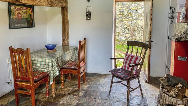 A rustic dining area with a checkered tablecloth, wooden chairs, a stone wall, and a view of greenery outside. - Birkerthwaire Stable-W41477 (Eskdale)