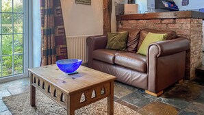 A cozy living room with a brown leather sofa, a wooden coffee table with a blue glass bowl, and a view of greenery through a large window. - Birkerthwaire Stable-W41477 (Eskdale)