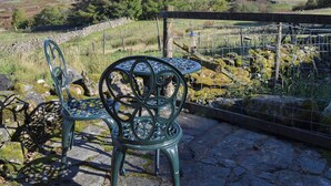 A rustic outdoor seating area with two metal chairs and a table, surrounded by stone walls and a wire fence, overlooking a mountainous landscape. - Birkerthwaire Stable-W41477 (Eskdale)
