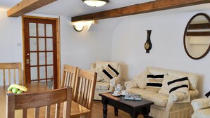 Een gezellige woonkamer met een houten eettafel, twee houten stoelen, een crèmekleurige bank met zwarte strepen, een klein bijzettafeltje en een wandklok.