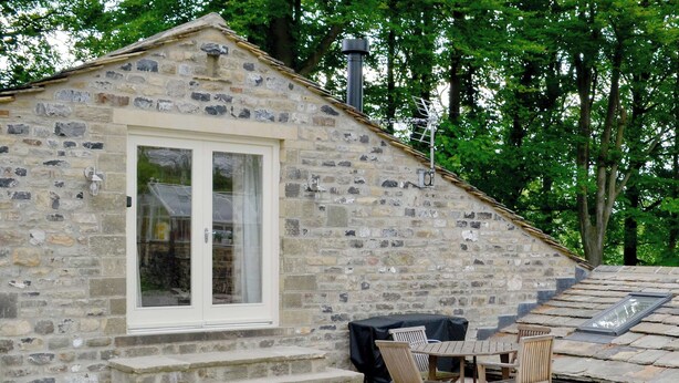 Un bâtiment en pierre avec une porte et des fenêtres blanches, un petit espace extérieur aménagé avec une table et des chaises, et une cheminée.