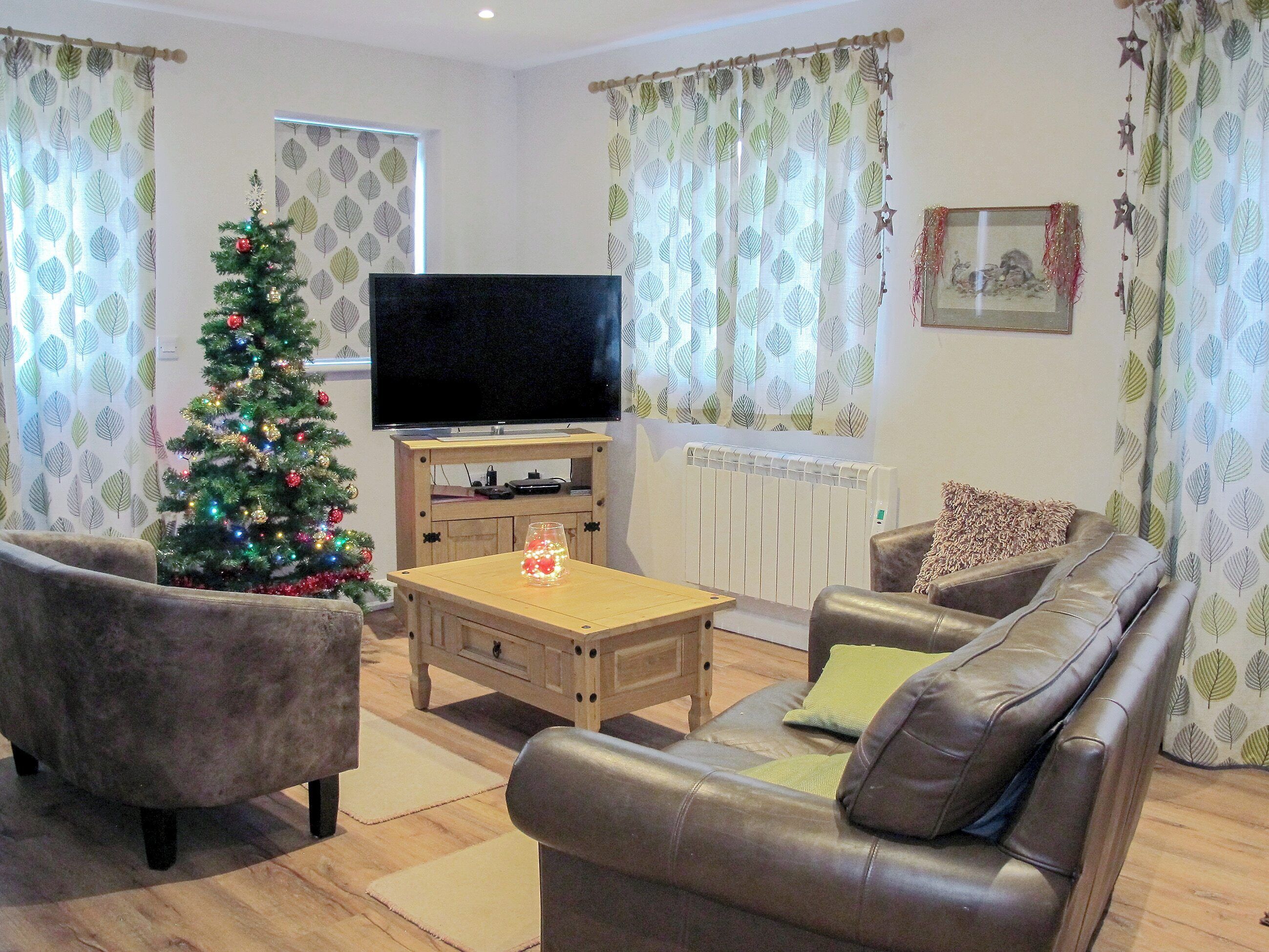 A living room with a Christmas tree, leather sofas, a flat-screen TV, and patterned curtains.