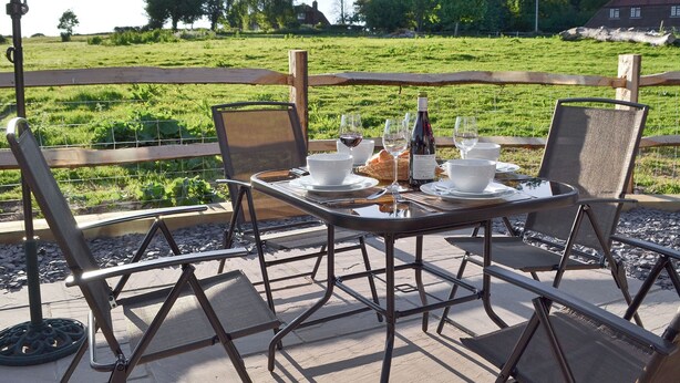 Un espacio de comedor al aire libre con una mesa dispuesta para cuatro, rodeada de sillas de metal, con vista a un campo cubierto de césped, árboles y una casa al fondo.