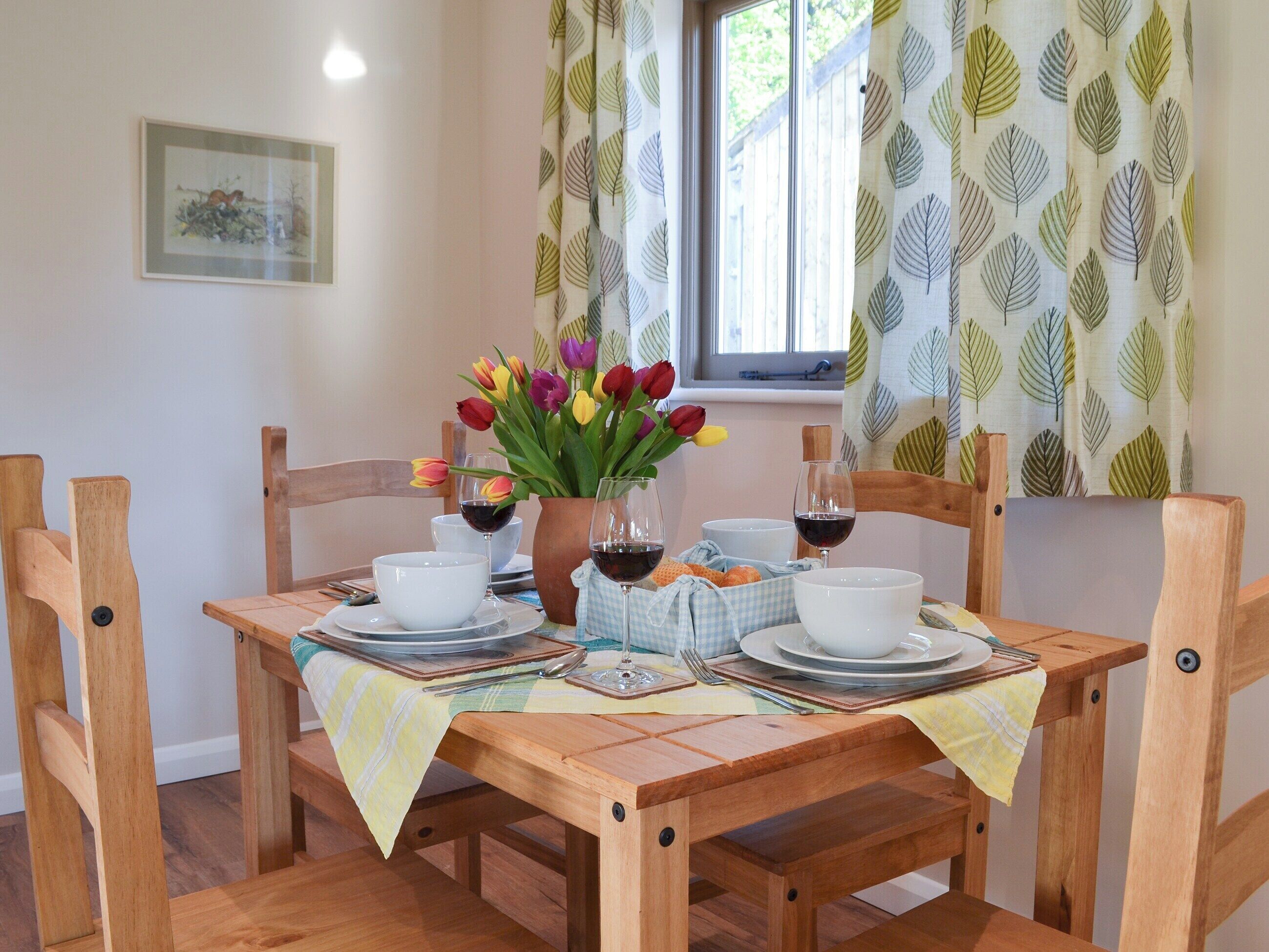 A dining table set with white plates, bowls, and glasses, surrounded by wooden chairs. A vase with colorful tulips and a bottle of red wine are on the table.