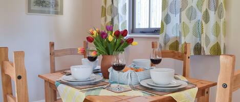 A dining table set with white plates, bowls, and glasses, surrounded by wooden chairs. A vase with colorful tulips and a bottle of red wine are on the table.