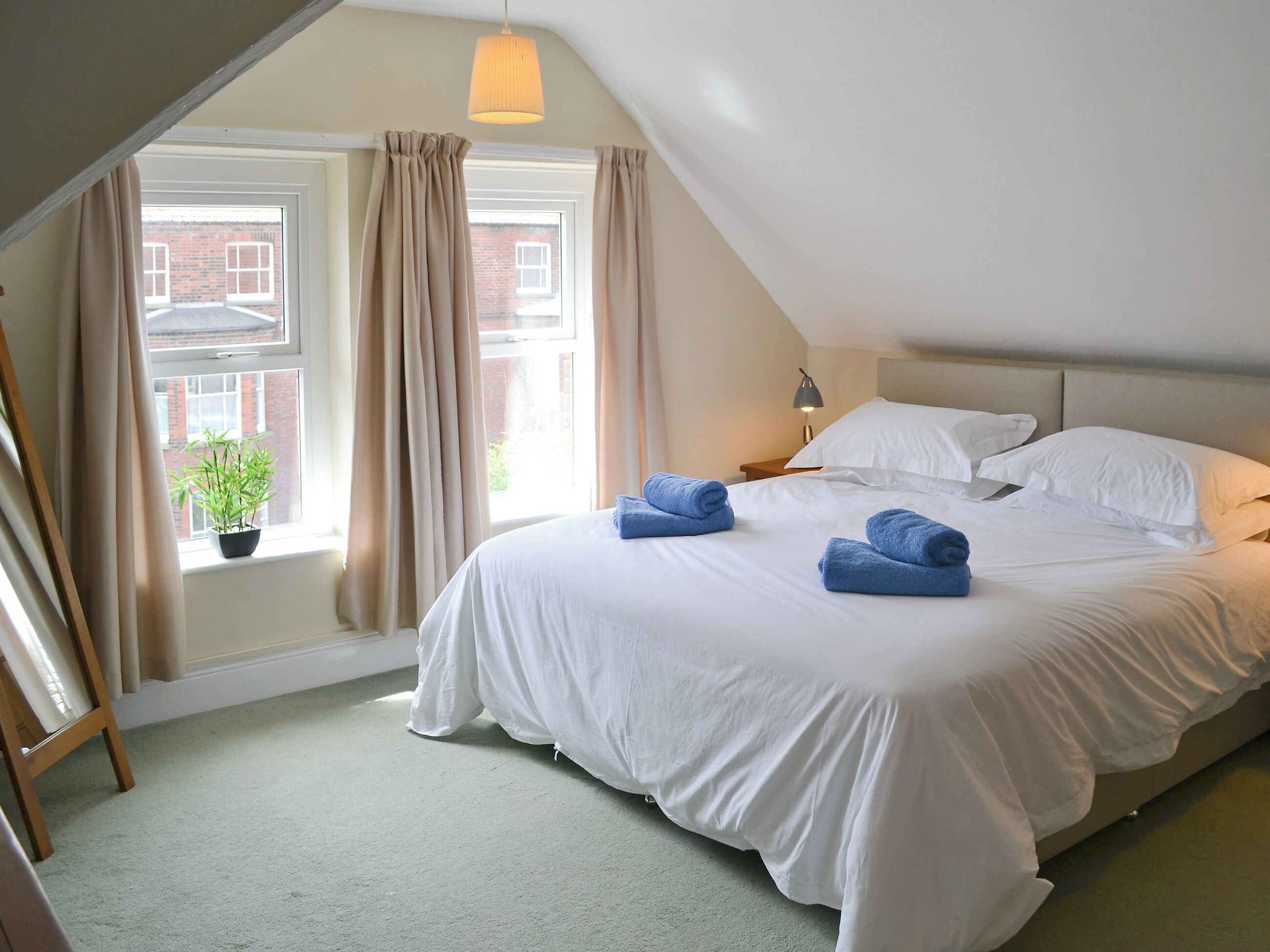 A bedroom with a large bed, a window with curtains, a small potted plant, and a lamp on a bedside table.