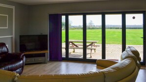 A living room with a television, a sofa, and a large sliding glass door leading to an outdoor area with a picnic table. - Aimmee's Lodge - UKC2203 (Doddington, near Lincoln)