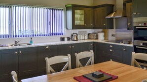 A modern kitchen with dark cabinetry, a wooden dining table, and a window with blinds. - Aimmee's Lodge - UKC2203 (Doddington, near Lincoln)