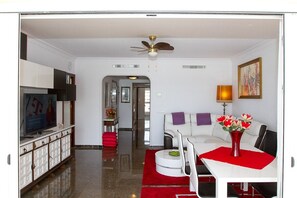 A modern living room with a flat-screen TV, a white sofa, a dining table with chairs, and a red rug.