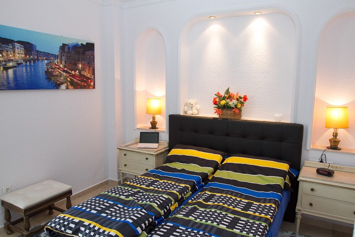 Un dormitorio con cama, mesitas de noche, lámpara, un osito de peluche y un cuadro de un paisaje urbano.