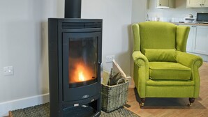 A modern living room with a black wood-burning stove, a green armchair, and a kitchen area in the background.