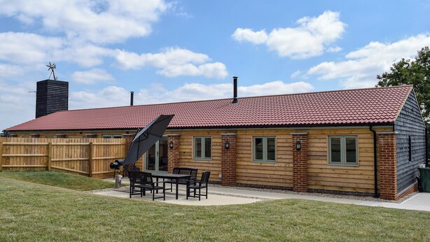 A wooden cabin with a red-tiled roof, a patio with a table and chairs, and a fenced yard.
