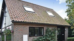 A two-story brick house with a tiled roof, two skylights, a black door, and a small garden area with a hedge and flowering plants. - Broad Cottage Boathouse (Ranworth, near Wroxham)