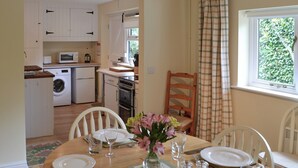 A dining area with a table set for four, a kitchen in the background, and a window with a view of greenery. - Broad Cottage Boathouse (Ranworth, near Wroxham)