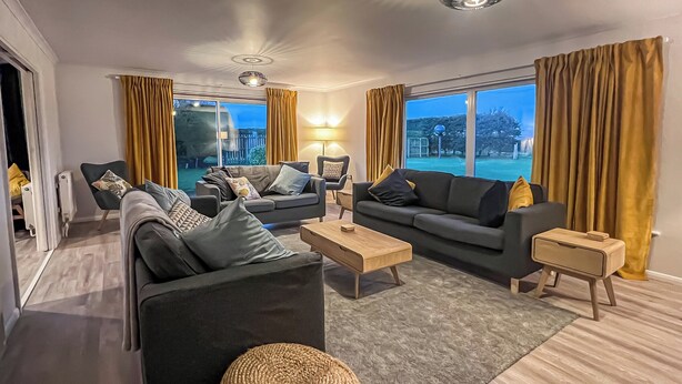 A modern living room with two sofas, a coffee table, and a side table. There are curtains on the windows, and a view of the outdoors is visible.