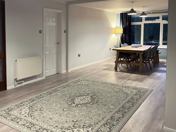 A spacious room with a large wooden dining table, a patterned rug, and a view of the outdoors through a window.
