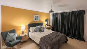 A bedroom with a bed, bedside tables, a chair, a desk, a lamp, a ceiling fan, and a window with curtains.