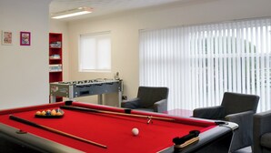 A pool table with balls, a foosball table, and two chairs in a room with a window and blinds.