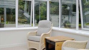 A cozy room with a wicker chair and a small table, large windows with a view of greenery outside.