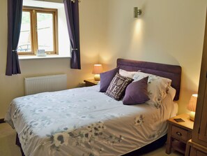 A bedroom with a bed, two bedside tables, a window with curtains, a wall-mounted light fixture, and a radiator.