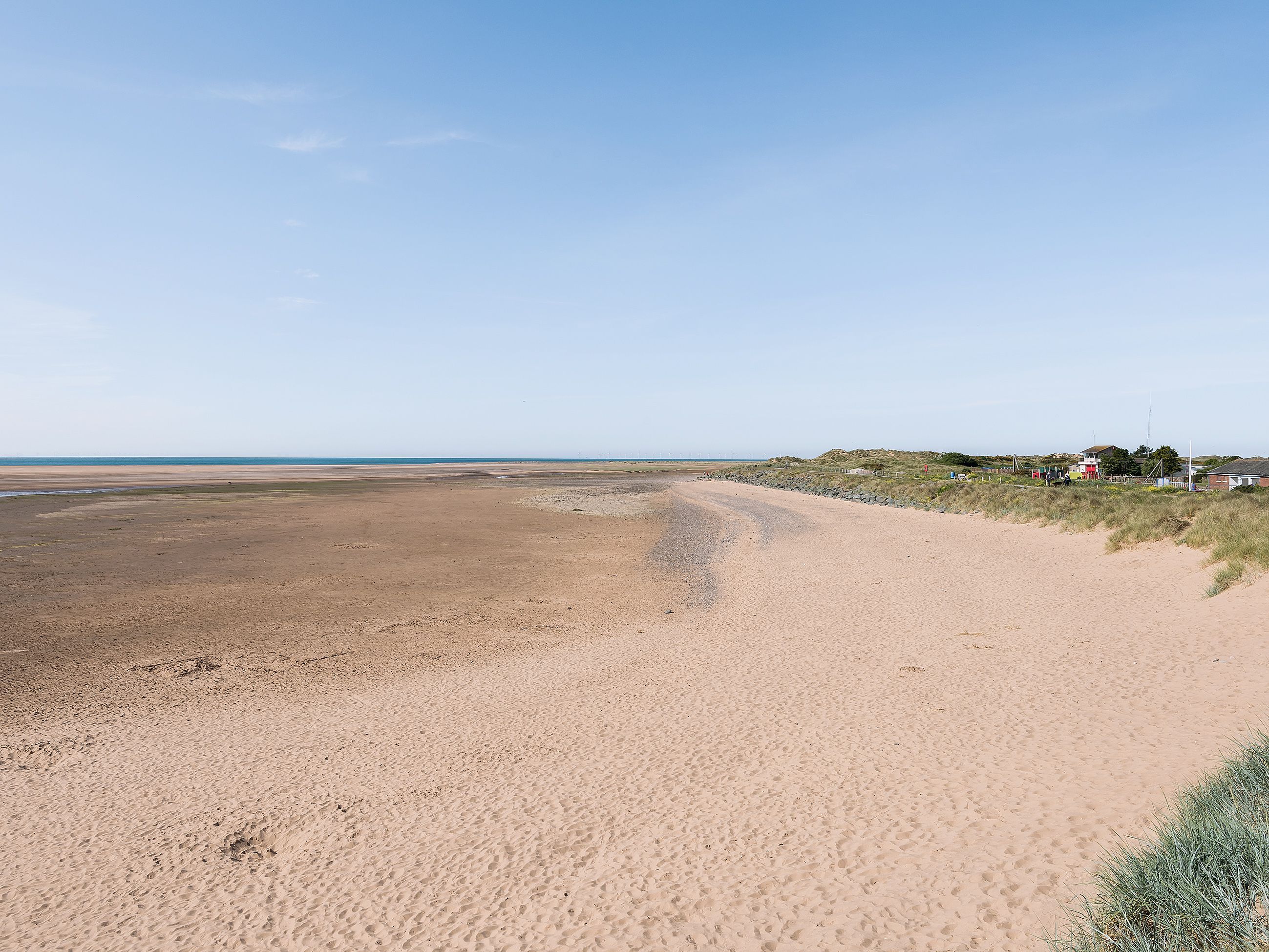Une plage de sable fin, un ciel dégagé, quelques bâtiments visibles au loin et une zone herbeuse délimitée par une clôture.